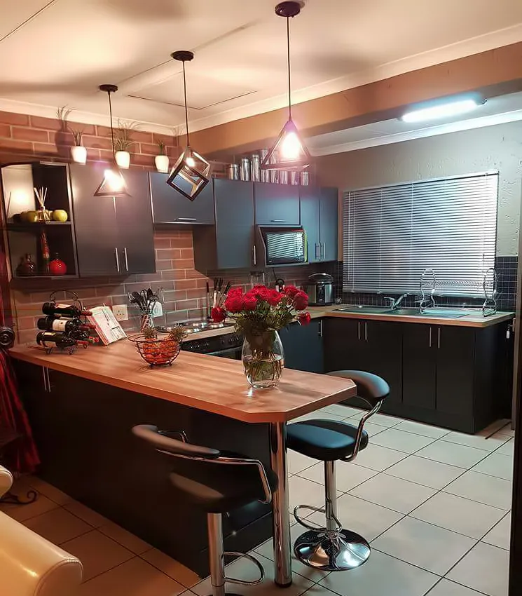 After: the kitchen with charcoal cabinets, a brick-look wall, a timber breakfast bar with two bar stools and three geometric pendant lights.
