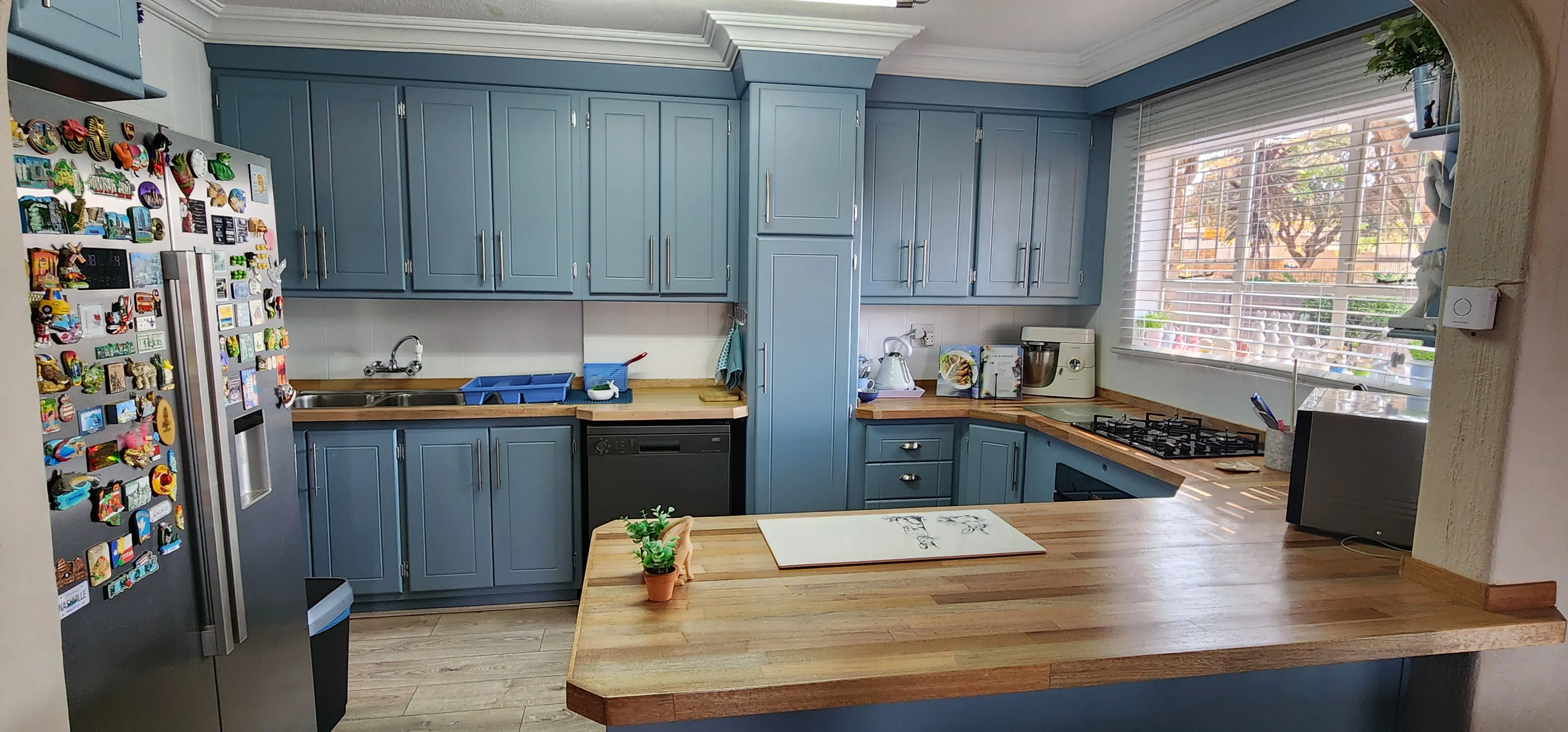After: the kitchen with dusty blue cabinets, timber countertops and white blinds at the window.