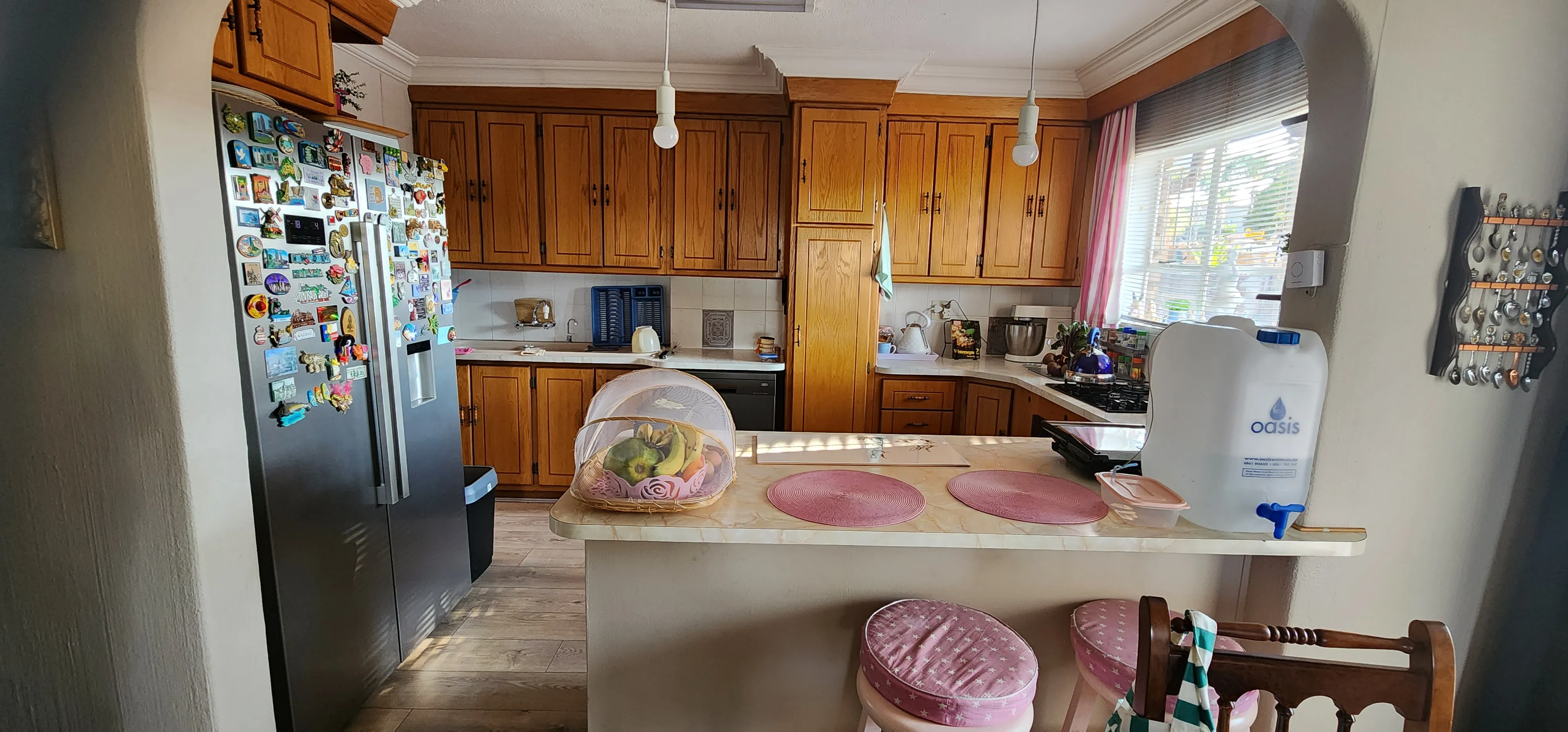 Before: a kitchen seen through an archway, with honey-coloured timber cabinets, a pale breakfast counter and a fridge covered in magnets.