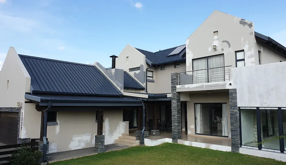 Before: a double-storey gabled house with beige walls marked by pale repair patches, a dark metal roof and stacked-stone columns.