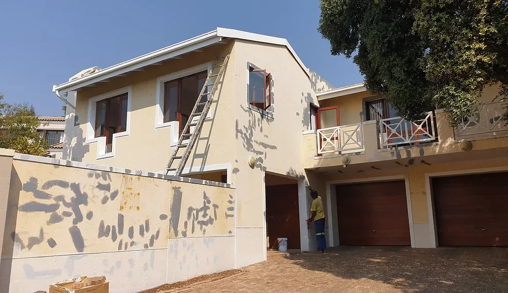 Before: a double-storey house with pale yellow walls covered in grey repair patches, a ladder against the wall and a painter at the entrance.