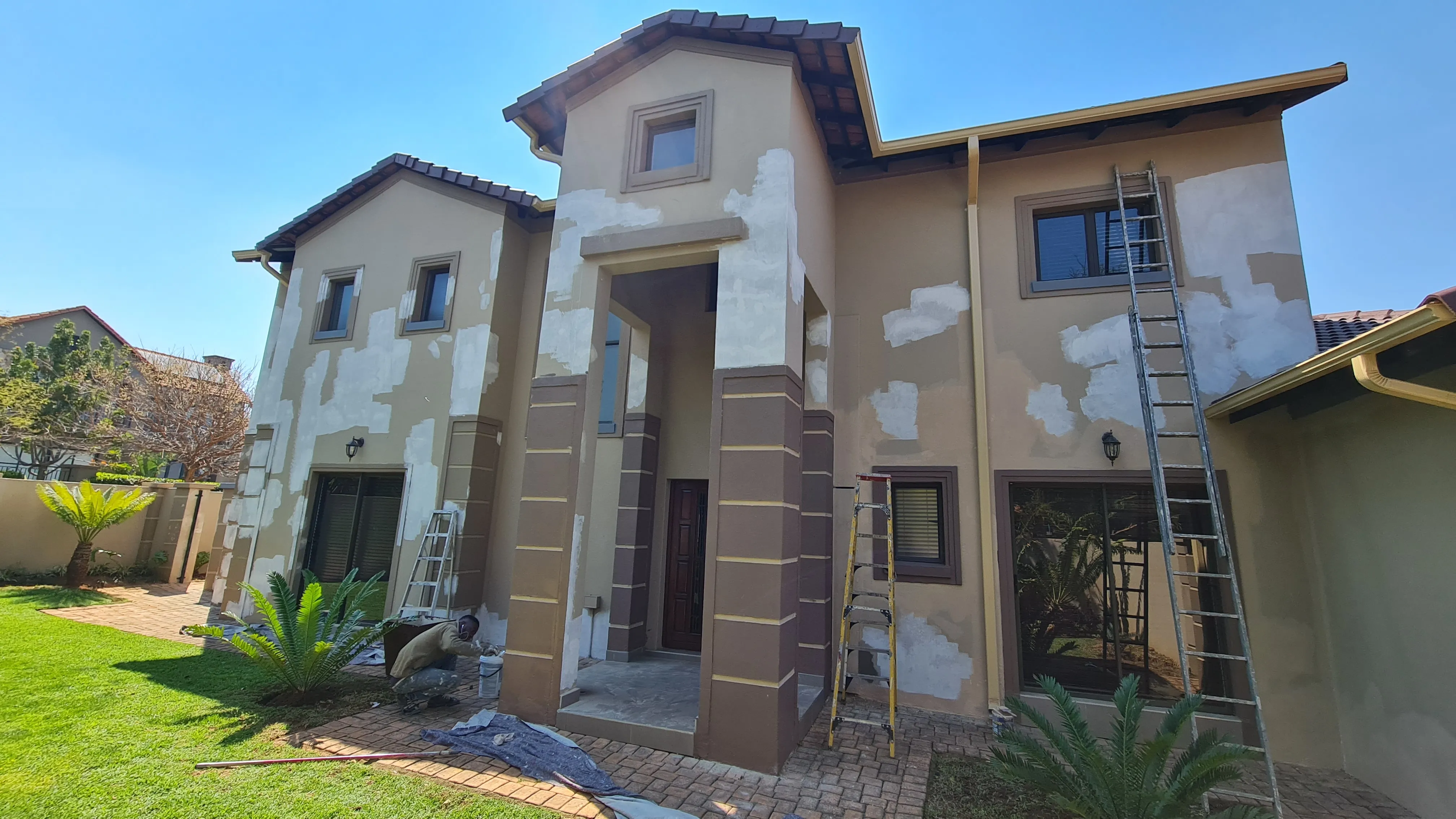 Before: a double-storey house with tan walls covered in white repair patches, ladders against the facade and a painter at work.