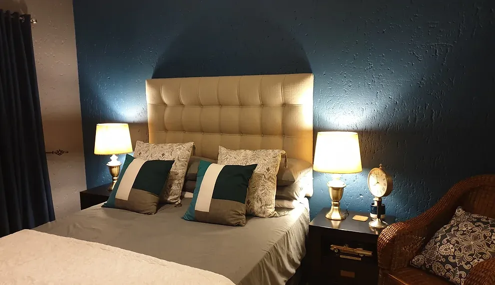 After: the bedroom with a deep teal wall, a cream buttoned headboard, brass bedside lamps and teal cushions.