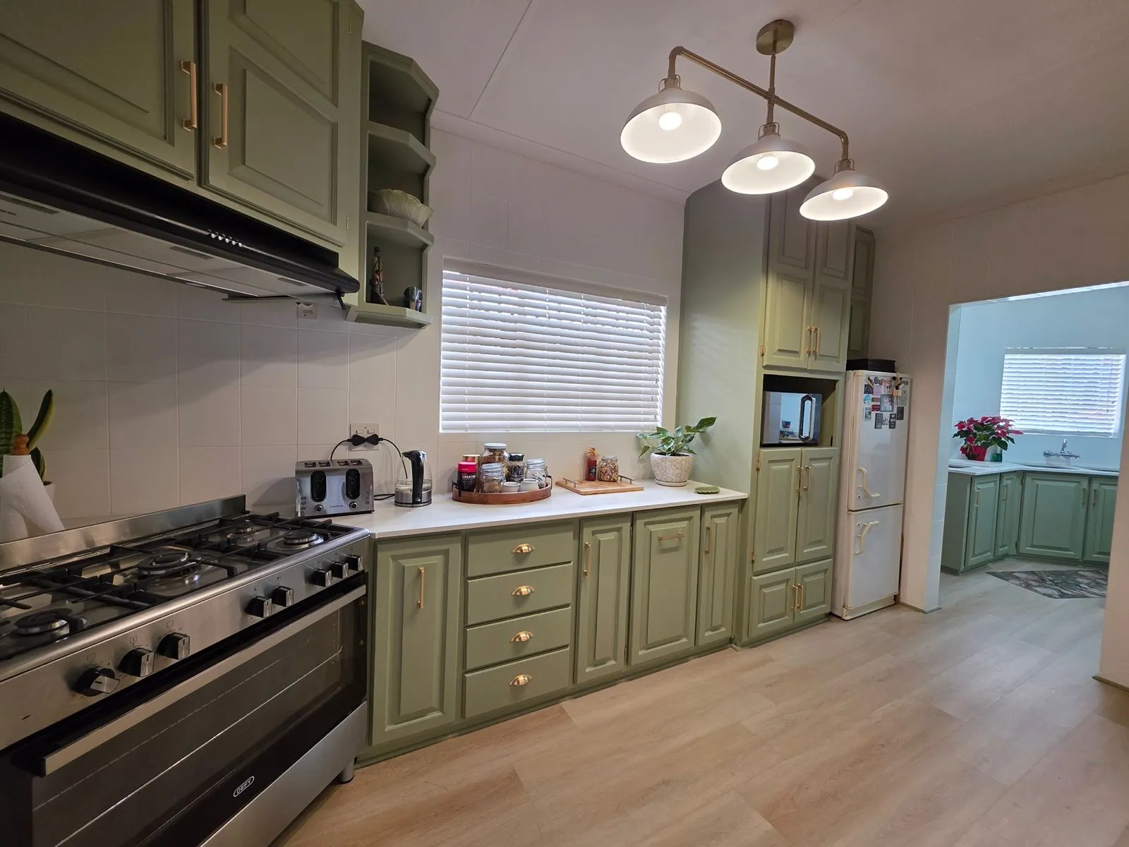 After: sage green cabinets with brass-toned handles, white countertops, a freestanding range cooker and a three-shade pendant light.