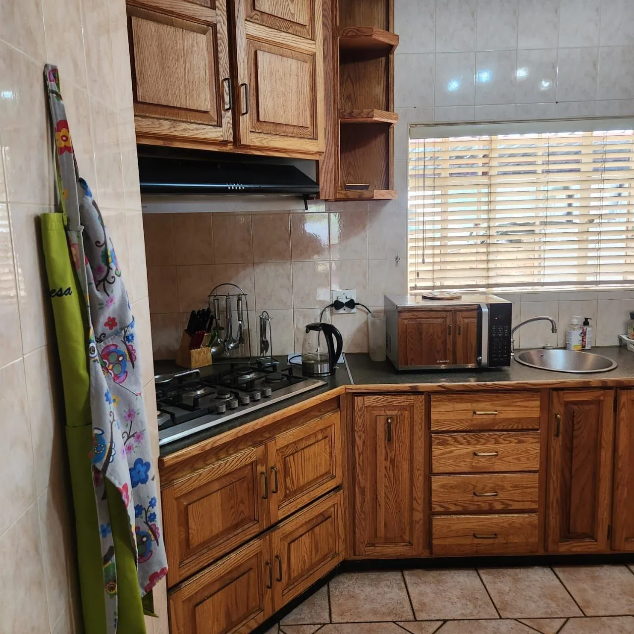 Before: oak kitchen cabinets around a corner gas hob, with beige wall tiles and a window with timber blinds.
