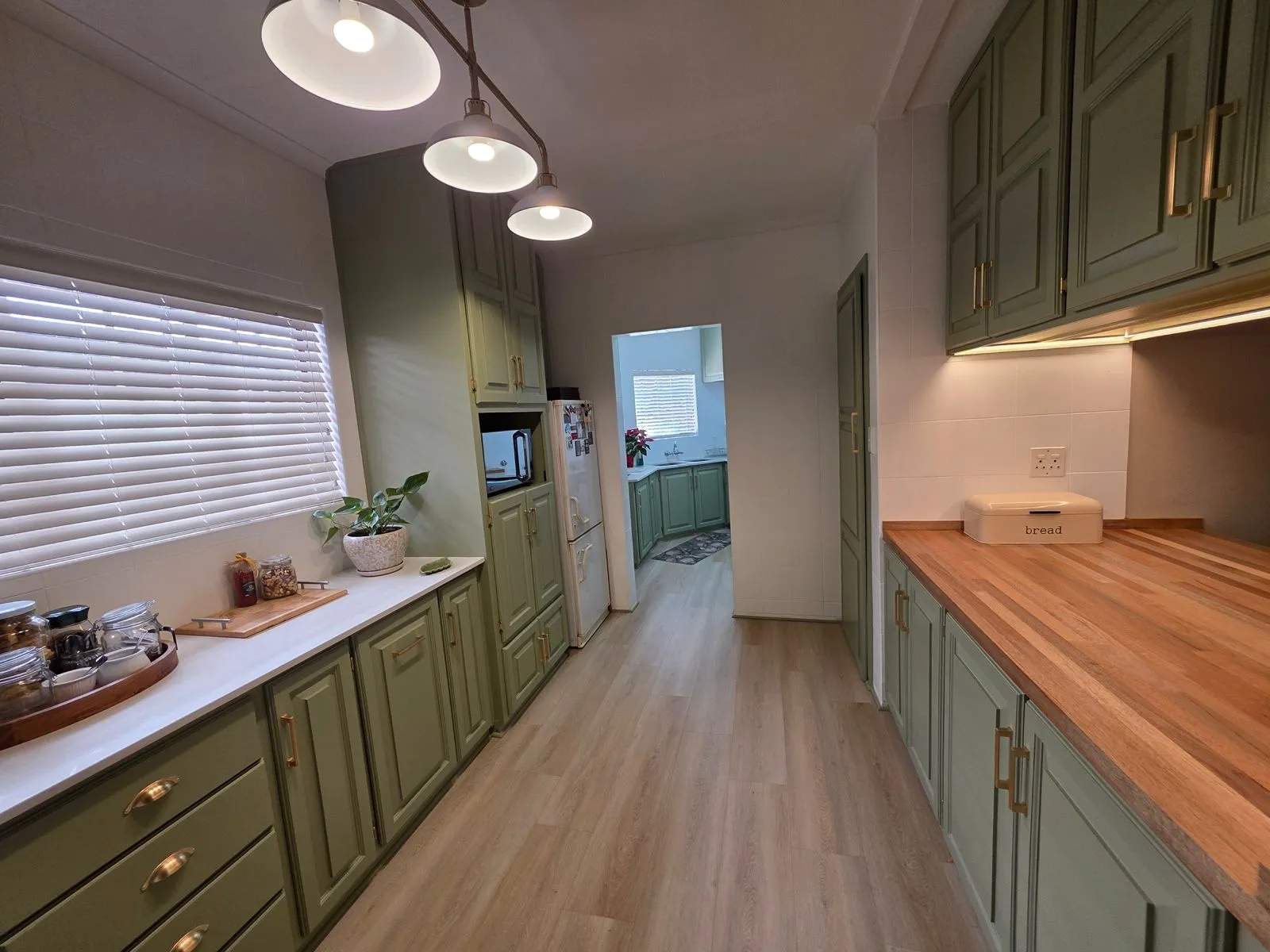 After: the same run of cabinets in sage green beneath a three-shade pendant, a timber countertop opposite and the scullery beyond.