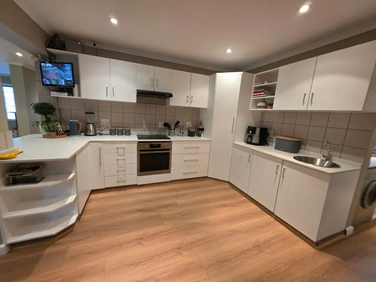 After: the same kitchen with white cabinets and countertops, a taupe tiled splashback and timber-look flooring.