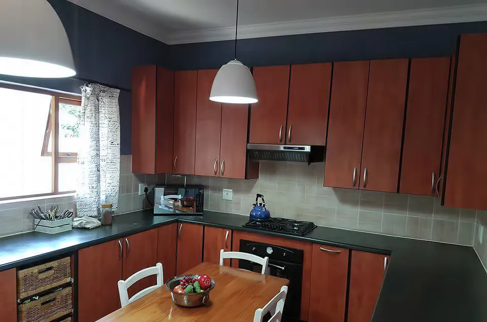 Before: a kitchen with cherry-brown cabinets, a navy wall above, beige splashback tiles and a pine table in the foreground.